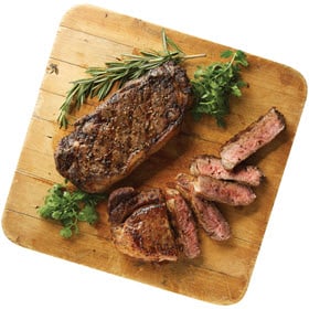 Two Boneless NY Strip Steak from Dierbergs shown grilled and resting on a wooden board, one sliced and ready to be served.