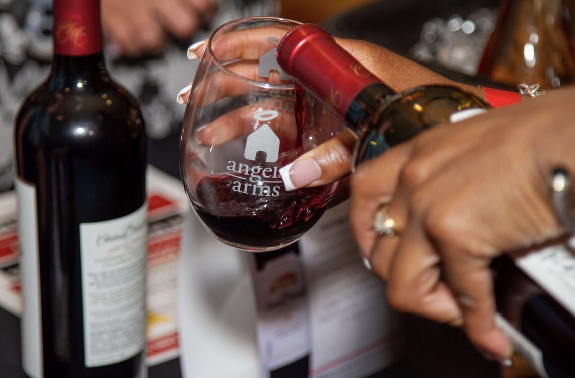 Close up on a woman's hand pouring wine into a stemless wine glass with the Angels' Arms logo on it. Other wine bottles are seen blurred in the background.