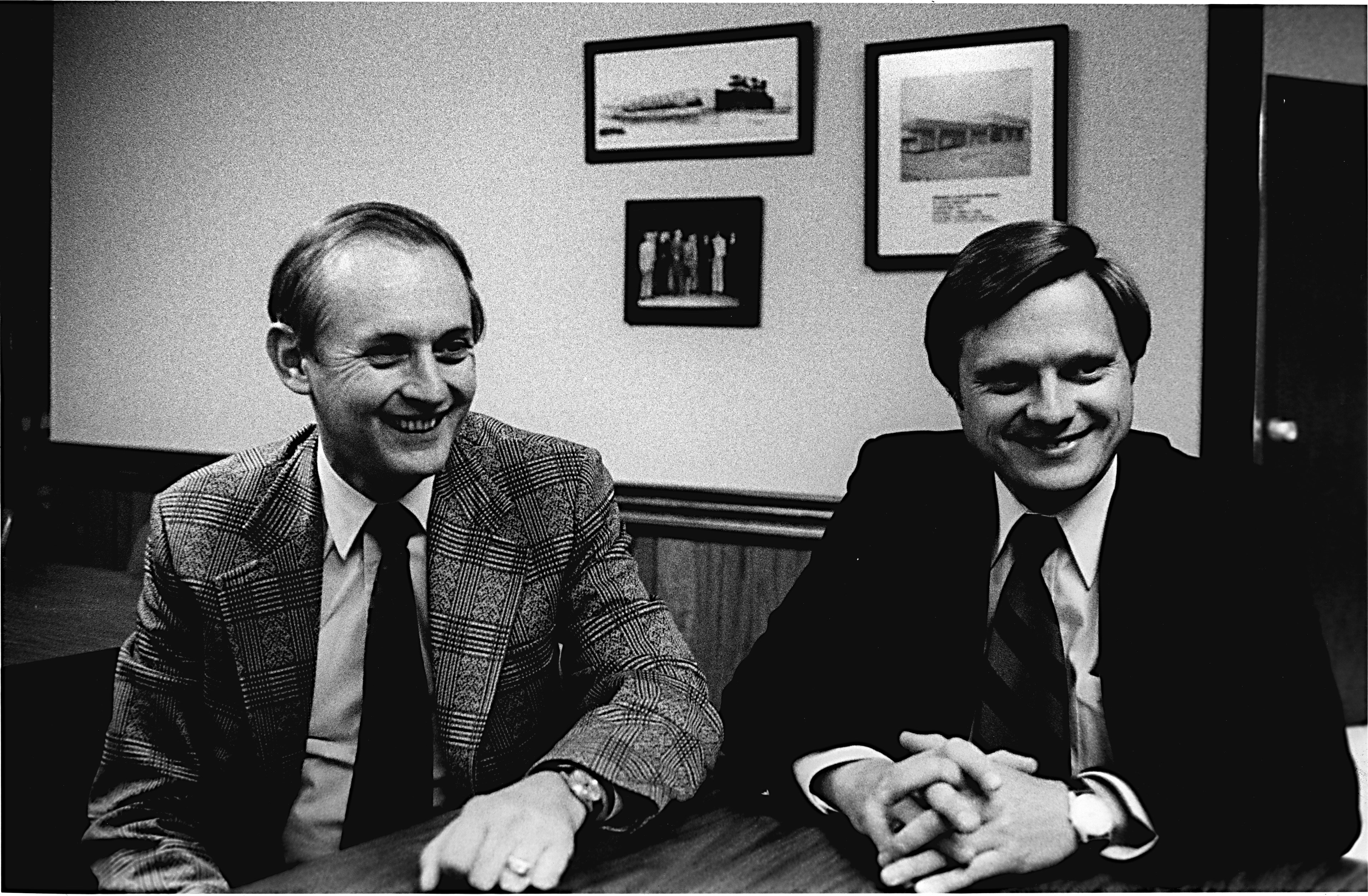 A photo of Roger Dierberg and Bob Dierberg circa 1977, sitting at a table in an office, both wearing suit jackets and ties, smiling as they speak to someone off camera.